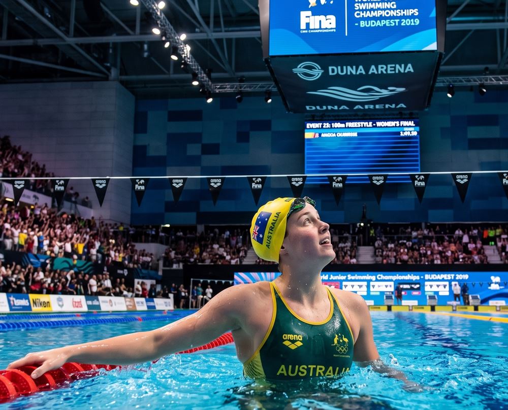 Joven nadadora australiana en la piscina de la Duna Arena, mirando el marcador oficial del 7º Campeonato Mundial Júnior de Natación FINA Budapest 2019, con las gradas llenas de espectadores al fondo.