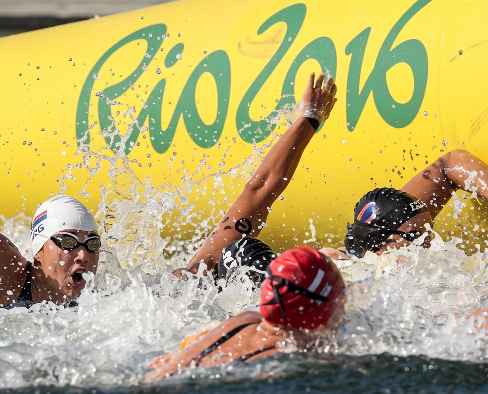Una fotografía de acción de varios nadadores olímpicos compitiendo en una carrera de natación en aguas abiertas durante los Juegos Olímpicos de Río 2016. Un nadador con un gorro blanco y gafas está en primer plano con la boca abierta, luchando a través de agua salpicada. Otros competidores con gorros negros, rojos y oscuros están cerca. Detrás de ellos, una gran boya inflable amarilla muestra el logotipo "Rio 2016" en verde y texto parcial "MARINHA DO
