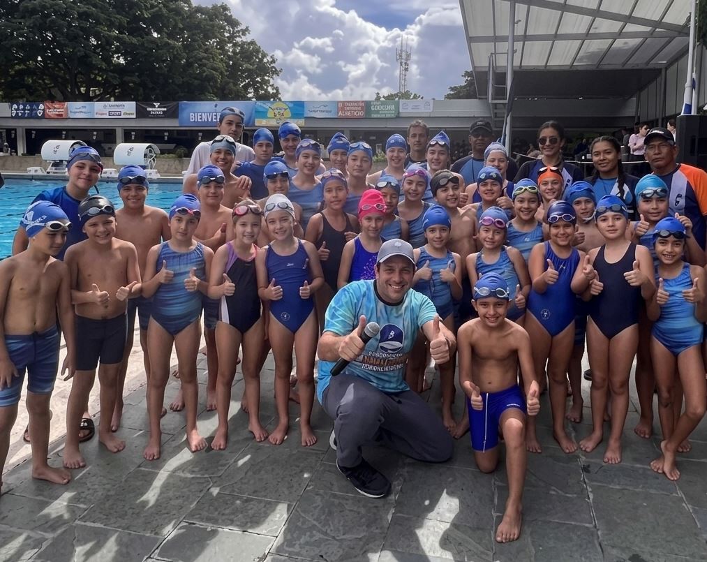 decenas de niños y niñas nadadores sonriendo y levantando los pulgares junto a una piscina olímpica al aire libre. En el centro, un hombre adulto arrodillado sostiene un micrófono y posa alegremente, rodeado por los jóvenes atletas con gorros y trajes de baño azules durante la Copa Danny Chocrón.