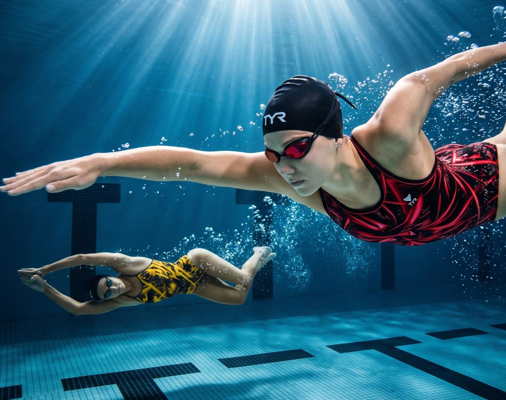 Una fotografía subacuática de alta resolución y ultra nítida de dos nadadoras profesionales en una piscina de competición. En primer plano a la derecha, una nadadora con gorro negro, gafas rojas y un traje de baño con estampado geométrico rojo y negro se desliza por el agua cristalina. En el fondo, a la izquierda, otra nadadora con traje de baño amarillo y negro sigue el ritmo. El fondo muestra mosaicos de piscina nítidos y la línea 'T' negra de carril, con haces de luz dramáticos que penetran desde arriba.