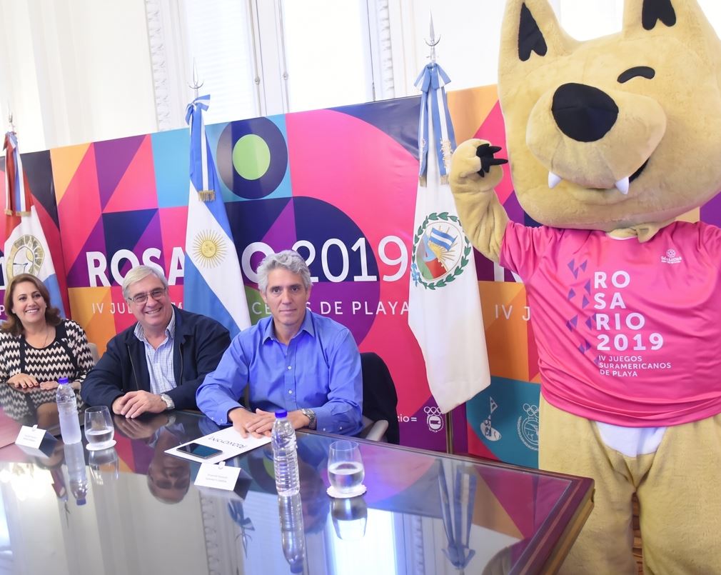 cinco autoridades sentadas detrás de una mesa de cristal durante una conferencia de prensa o evento oficial. De fondo, hay un panel colorido con el texto "Rosario 2019, IV Juegos Suramericanos de Playa" junto a banderas oficiales, incluyendo la bandera de Argentina. A la derecha de la imagen, de pie, posa animadamente la mascota del evento, una figura de un animal color arena que viste una camiseta deportiva de color rosa.