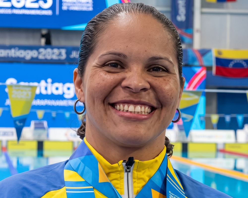 Belkis Mota, nadadora paralímpica venezolana S12, sonríe sosteniendo su medalla de plata en los Juegos Parapanamericanos Santiago 2023 frente a la piscina.