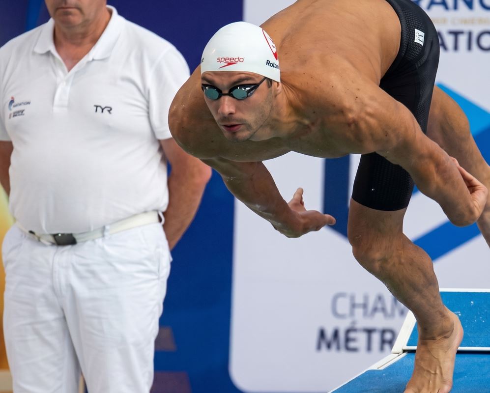 El nadador francés Roman Fuchs realizando una salida explosiva desde el poyete de salida azul en una competición oficial de natación.