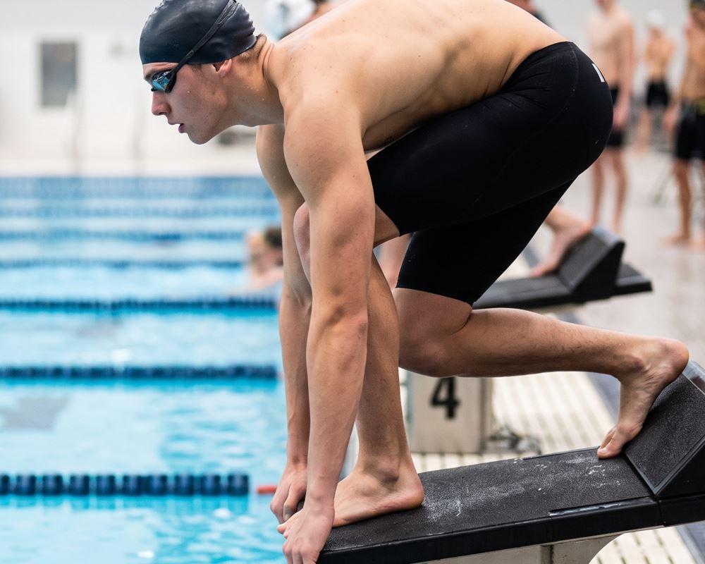 Nadador competitivo en posición "Take your marks" en el poyete, concentrado en su ejercicio para mejorar la velocidad de reacción auditiva y explosiva.