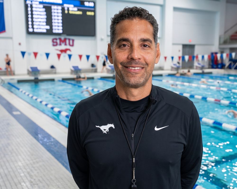 Retrato de Ozzie Quevedo, ex nadador venezolano, en su actual rol como entrenador de natación de SMU posando frente a una piscina de competición.