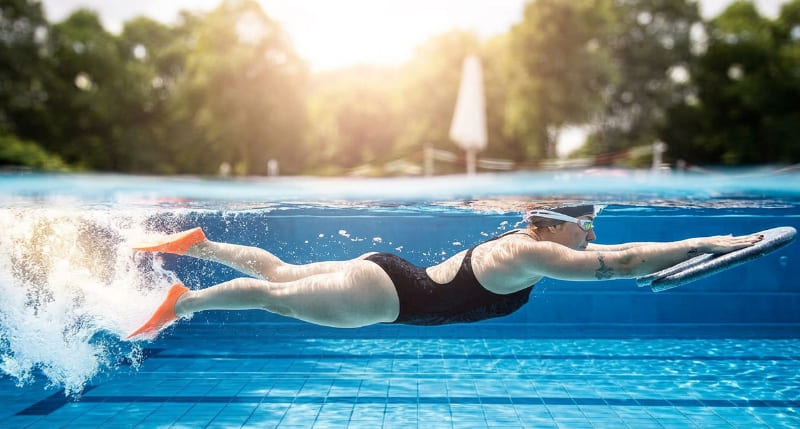 nadadora ejecutando una posición horizontal perfecta (streamline) bajo el agua en una piscina. Ella está sosteniendo una tabla de entrenamiento y usando aletas naranjas para potenciar el deslizamiento, demostrando una excelente
