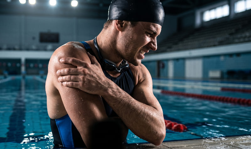 Nadador sujetándose el hombro con gesto de dolor al borde de una piscina olímpica, representando los síntomas del síndrome de hombro de nadador.